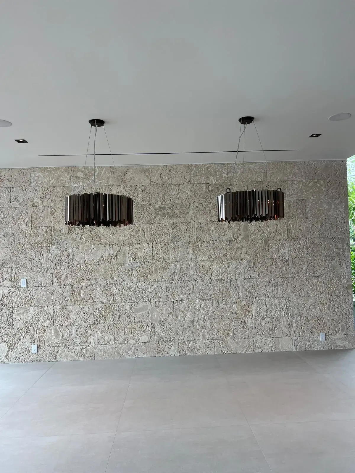 Two bronze drum pendant fixtures hanging from a white ceiling in front of a coral stone wall