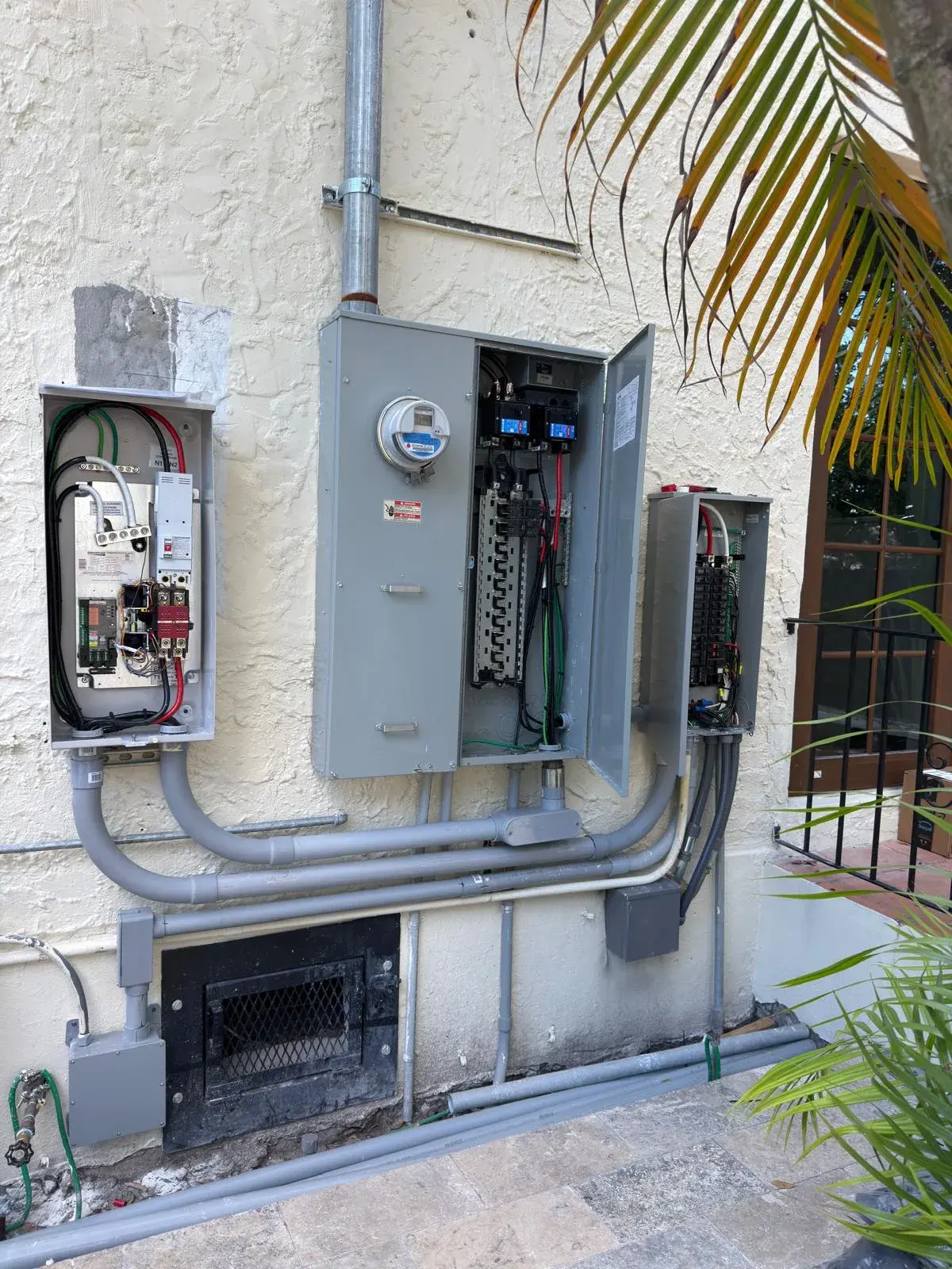 Stucco wall with an open transfer switch, a meter and main panel and a subpanel connected by gray conduit